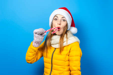 Young woman with a surprised face in a yellow jacket and hat of Santa Claus holds a candy cane on a...