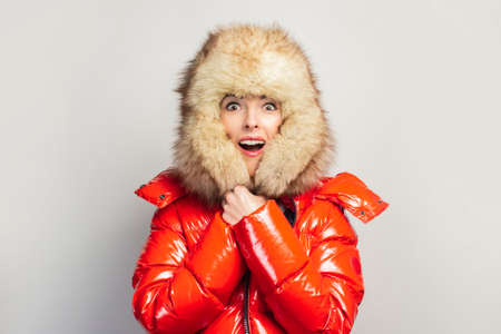 Surprised young girl in a red jacket and a fur hat, knits a hat on a light background. Concept shock, surprise.の写真素材