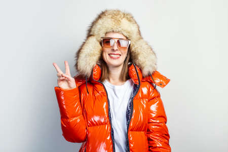 cheerful young girl in a red jacket, fur hat and sunglasses, shows the symbol of peace with v-sign with fingers on a light background.の写真素材