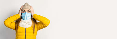young girl in a yellow jacket and hat with a medical mask on her face holding hands to her head on a...