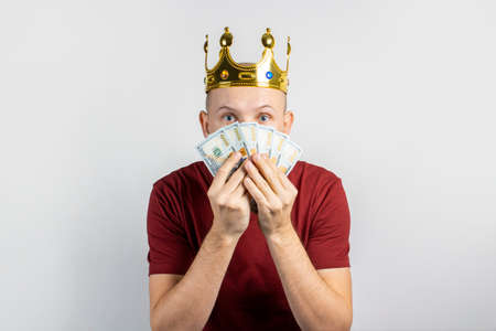 Young man in a golden crown with a surprised face holds money in his hands on a light background....