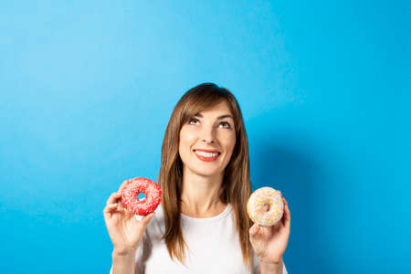 happy young girl in a white t-shirt holding two donuts on a blue background. Banner. Good mood, diet concept, bright colors.の写真素材