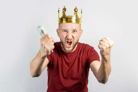 Young man in a golden crown holds money in his hands celebrates victory on a light background....