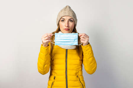 young girl in a yellow jacket and hat, holds a medical mask on a light background. Concept of the common cold, virus, infectious diseases.の写真素材