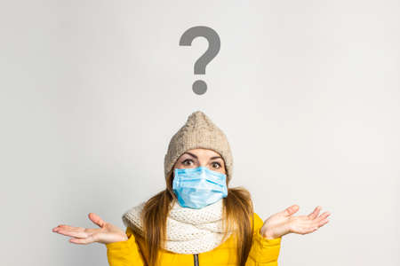 young girl in a yellow jacket and hat with a medical mask on her face throws up her hands, does not know what to do on a light background. Concept of the common cold, virus, infectious diseases.の写真素材