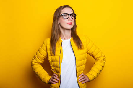 Friendly young woman in glasses and a yellow jacket looks to the side on a yellow backgroundの写真素材