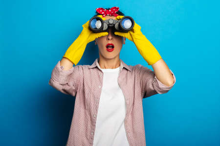 Surprised young woman in cleaning gloves looking through binoculars on blue backgroundの写真素材
