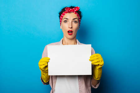 Surprised young woman in a headband, cleaning gloves holding a blank sheet on blue background