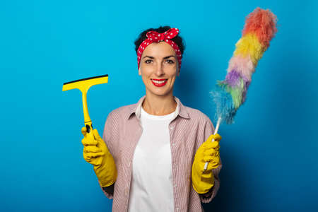 Smiling young woman in gloves holding glass scraper and dust brush on blue backgroundの写真素材