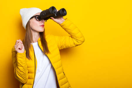 Young woman in yellow jacket looking through binoculars on yellow backgroundの写真素材