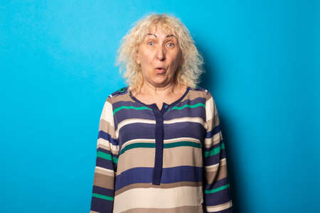 Surprised blonde curly old woman in striped dress on blue background