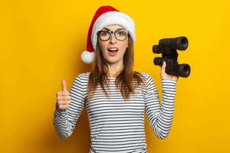 Young woman in a Santa Claus hat holds binoculars and makes a gesture All is well on a yellow...