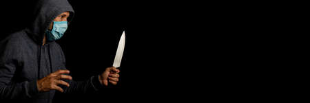 Aggressive young man in a medical mask with a knife on a black background. Bannerの写真素材