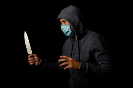 Aggressive young man in a medical mask in a hood with a knife on a black background
