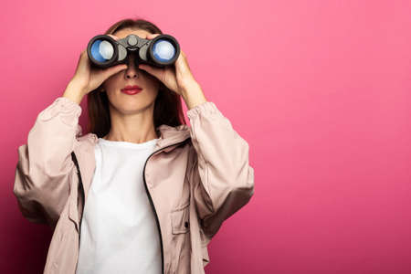 Young woman in jacket looking through binoculars on pink background