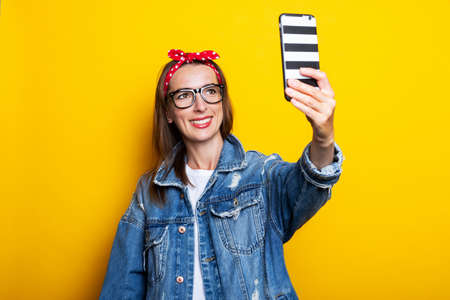 Young woman in denim jacket and glasses holds a phone in her hands and talks on video chat on a...