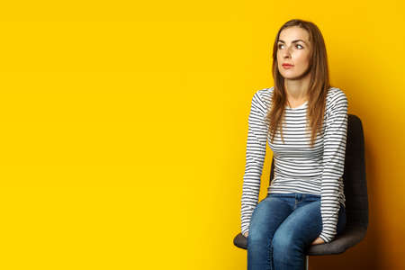Young woman with a smile sits on a chair and looks up on a yellow background. Banner.の写真素材