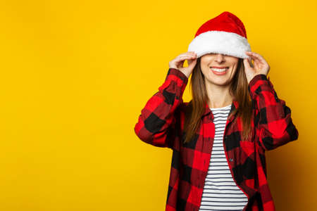 Young woman pulls santa hat and red plaid shirt over her eyes on yellow background.の写真素材