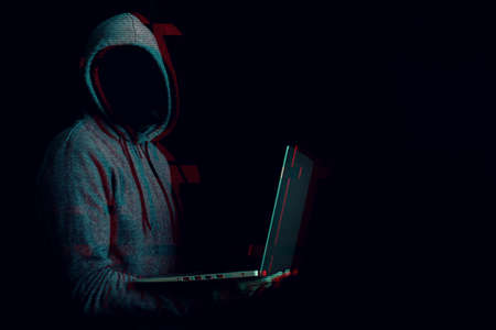 Man without a face in a hood holds a laptop in his hands on a dark background. Concept of cyber rogue, hacker. Banner. Added glitch effect.の写真素材