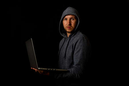 Man in a hood holds a laptop in his hands on a dark background. Concept of cyber rogue, hacker. Banner.の写真素材