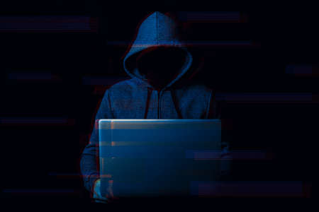 Man without a face in a hood holds a laptop in his hands on a dark background. Concept of cyber rogue, hacker. Banner. Added glitch effect.の写真素材