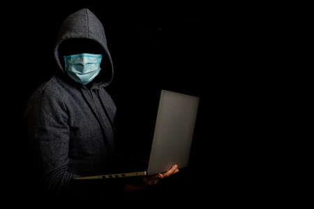 Man in a mask without a face and a hood holds a laptop in his hands on a dark background. Concept of cyber rogue, hacker. Banner.の写真素材