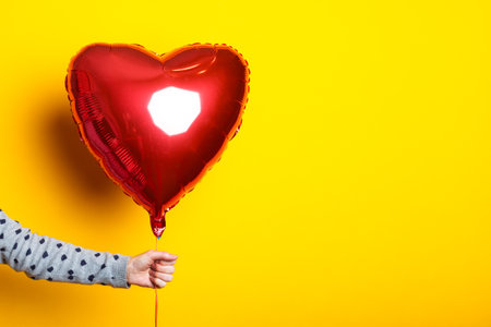 Female hand holds an inflatable heart-shaped balloon on a yellow backgroundの写真素材