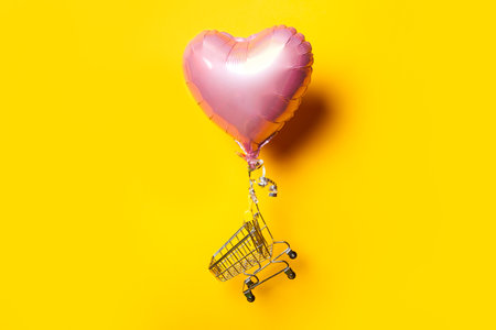 empty shopping trolley flies on pink heart shaped air balloon on yellow backgroundの写真素材