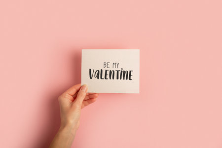Female hand holds a card on a pastel pink background. Valentine's day composition. Banner. Flat lay, top view.の写真素材