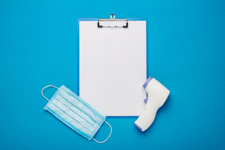 Digital non-contact thermometer, face shield and clipboard on a blue background. Banner. Top view, flat lay.の写真素材