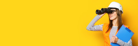 Young woman in signal vest and hard hat holds clipboard and looks through binoculars on yellow background.の写真素材