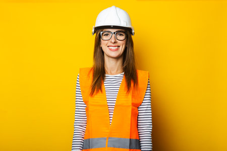 Young woman with a smile in a vest and hard hat on a yellow background. Construction concept, new...