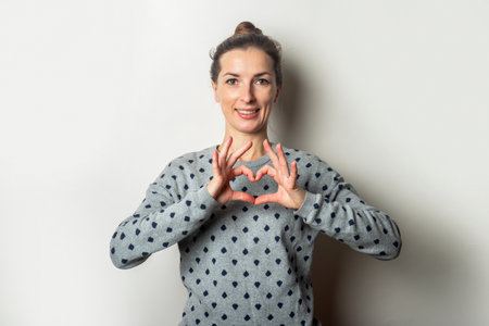 Young woman in a sweater makes a heart gesture on a light background. Valentine's Day concept....