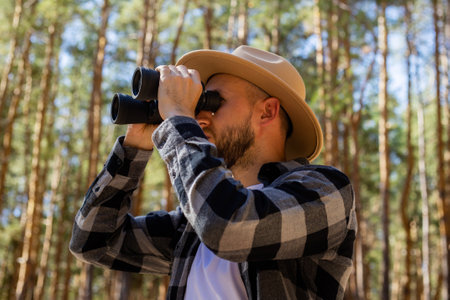 Men tourist in a hat and a gray checkered shirt looks through binoculars on a forest background.の写真素材
