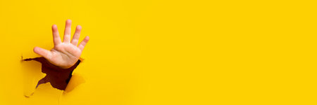 children's hand sticks out of a hole in a sheet of paper on a yellow background. Bannerの写真素材