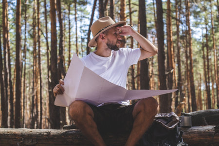 Bearded man in a hat holds a map in his hands during a halt on a hike. Hike in the mountains,...