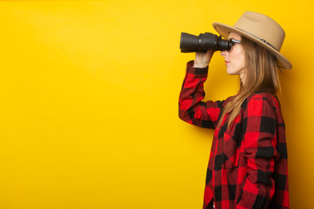 Young woman in hat and plaid shirt looking through binoculars on yellow background.の写真素材