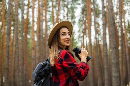 Young woman tourist with backpack, hat and red plaid shirt holding binoculars in the forest.