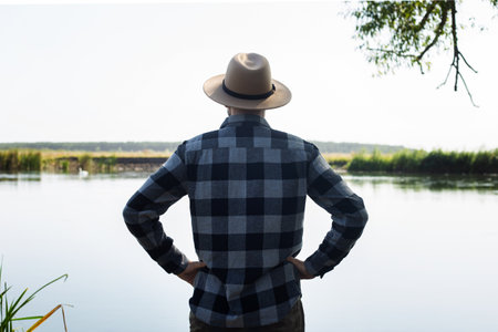 Man in a hat and a plaid shirt admires the landscape by the river.