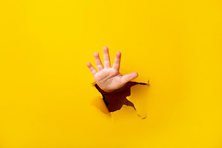 children's hand sticks out of a hole in a sheet of paper on a yellow background. Five finger sign,...
