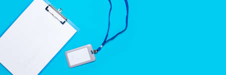 Clipboard with a blank sheet and a plastic name badge with blue lace on a light blue background....