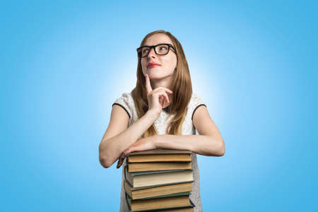 Young girl student with glasses leans on a pile of books and looks up with a pensive face on a blue...