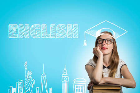 Young girl student with glasses leaning on a pile of books and looking up with a pensive face on a blue background with the text English. Concept of studying English and education abroad.の写真素材