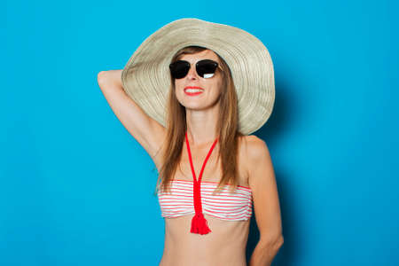 A young woman in a swimsuit, glasses and a wide-brimmed hat looks up on a blue background. Concept...