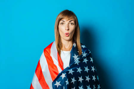 Young woman wrapped in USA flag with a surprised face on a blue background. USA visa concept, English language, gesture is all right, thumbs up, like.の写真素材