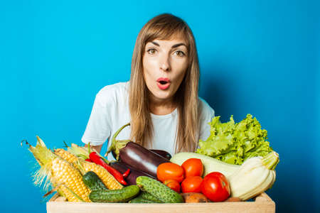 Young woman holds a box with fresh vegetables on a blue background. Good harvest concept, natural product.の写真素材