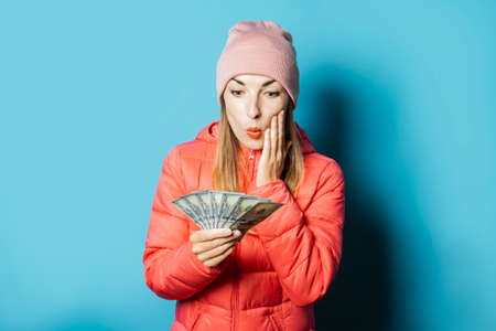 Beautiful young woman in a hat and a winter jacket with a surprised face holds money on a blue...