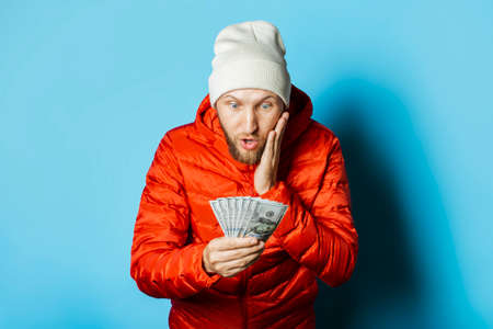 Man with a surprised face in a hat and a red jacket parka holds money in his hands on a blue...