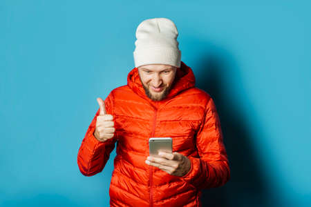Man with a happy face in a hat and a red jacket holds a mobile phone in his hands on a blue...
