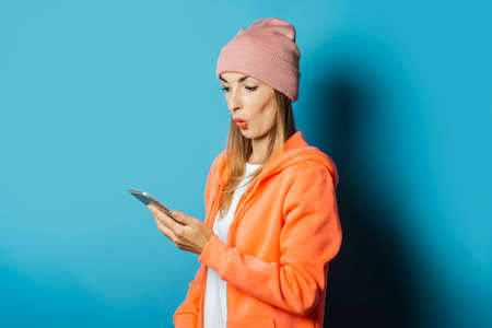 Beautiful Young woman in a hoodie and a hat with a surprised face holds a mobile phone on a blue...